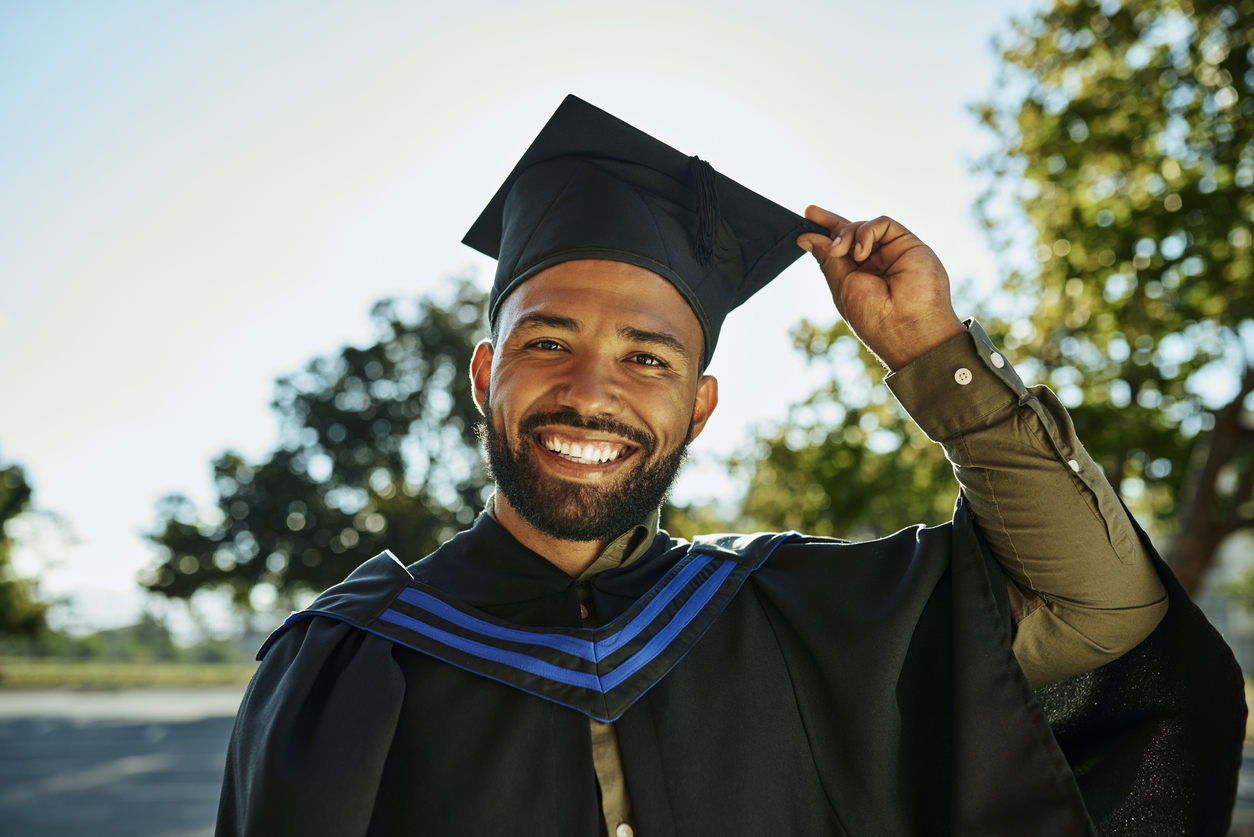 Portrait of smiling graduate in gown and hat standing alone on university campus at graduation ceremony. Excited, happy qualified postgrad graduating college for academic bachelor, masters and degree